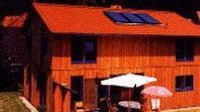 Ein modernes Holzhaus mit rotem Dach und Solarpanelen, umgeben von grüner Natur und einem einladenden Gartenbereich.