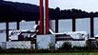 Das Bild zeigt eine moderne Fabrik mit großen Fenstern und zwei hohen roten Schornsteinen, eingebettet in eine ländliche Umgebung.