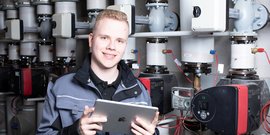 Ein junger Anlagenmechaniker vor einer Heizungsanlage, der ein Tablet hält 