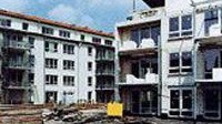 Ein Blick auf eine Baustelle mit zwei Wohnblöcken, die teilweise fertiggestellt sind, und einem leeren Platz im Vordergrund.