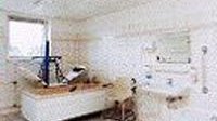Der Raum ist minimalistisch gestaltet, mit weißen Wänden und Fliesen, und enthält eine Liege sowie verschiedene medizinische Utensilien.