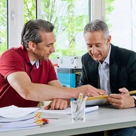 Zwei Männer sitzen an einem Tisch und diskutieren intensiv über Dokumente, während sie auf einen Bildschirm schauen.