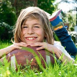 Ein fröhliches Kind liegt im Gras und hält einen Football, während die Sonne auf sein lächelndes Gesicht scheint.