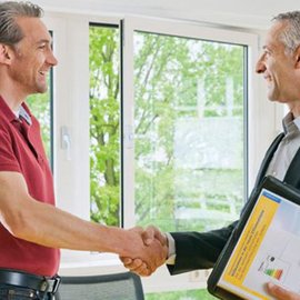 Zwei Männer schütteln sich die Hände in einem hellen Büro mit großen Fenstern und Blick auf grüne Bäume.