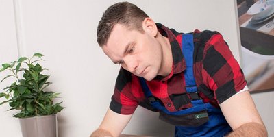 Ein Mann in einem karierten Hemd und blauer Latzhose konzentriert sich auf eine Aufgabe an einem Tisch.