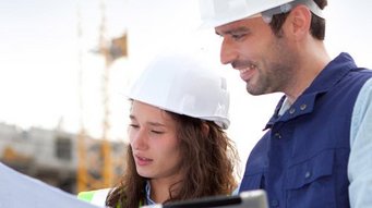 Eine Frau mit Bauhelm hält eine Zeichnung neben ihr steht ein Mann mit einem Tablet auf einer Baustelle